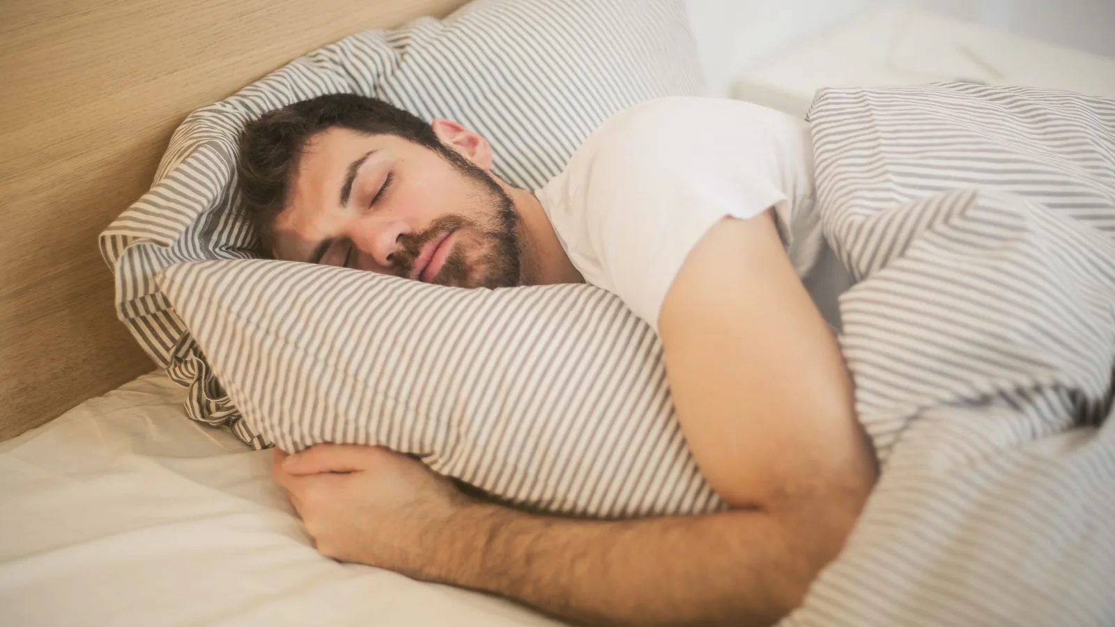 A man asleep in bed in soft morning light, one bare wrist resting on the duvet.