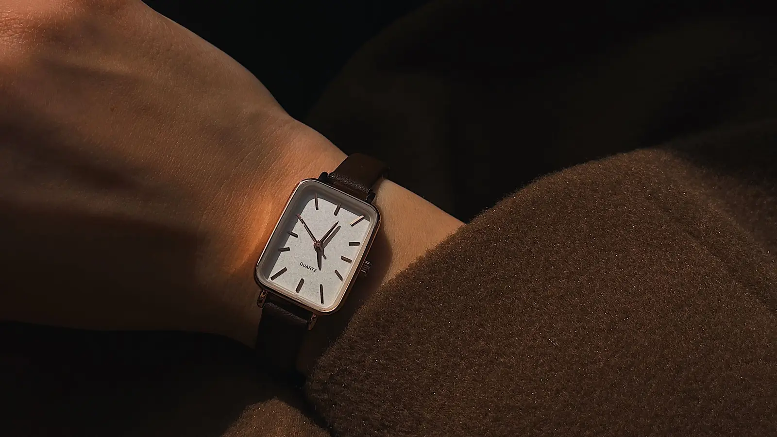 A simple analog watch on a wrist, lit by low afternoon sun against a wool coat.