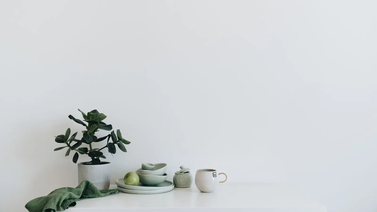A calm still life of sage ceramics, a pear and a plant on a white surface.