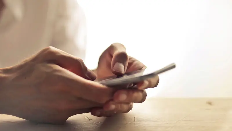 Two hands holding a phone on a wooden table, backlit by a bright window.
