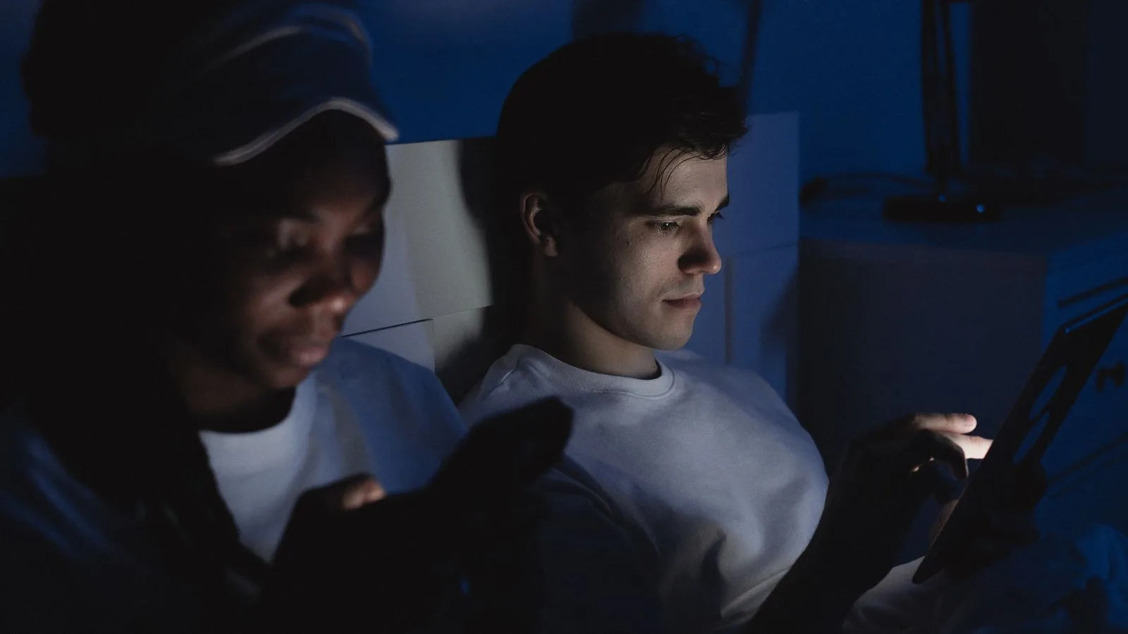 Two people lying in a dark bedroom, faces lit by the screens of their devices.