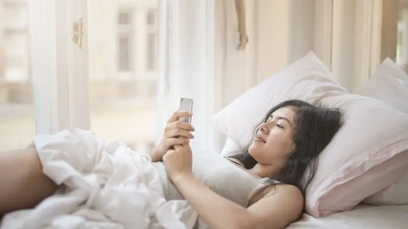A woman lying in bed in soft morning light, looking at her phone.
