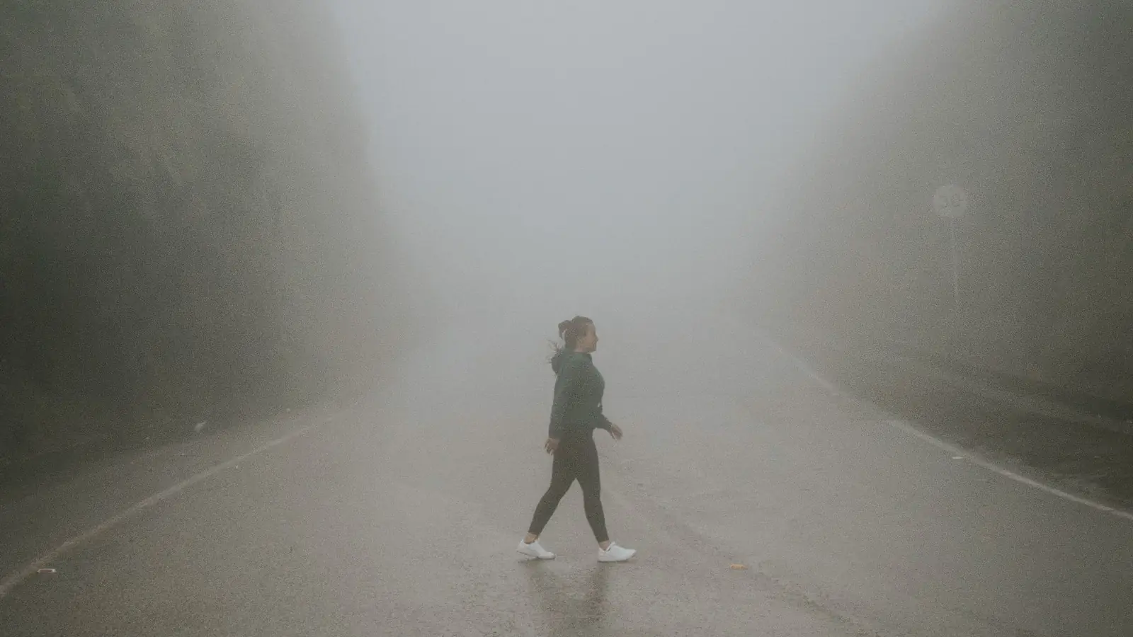 A person walking across an empty road in heavy morning mist.