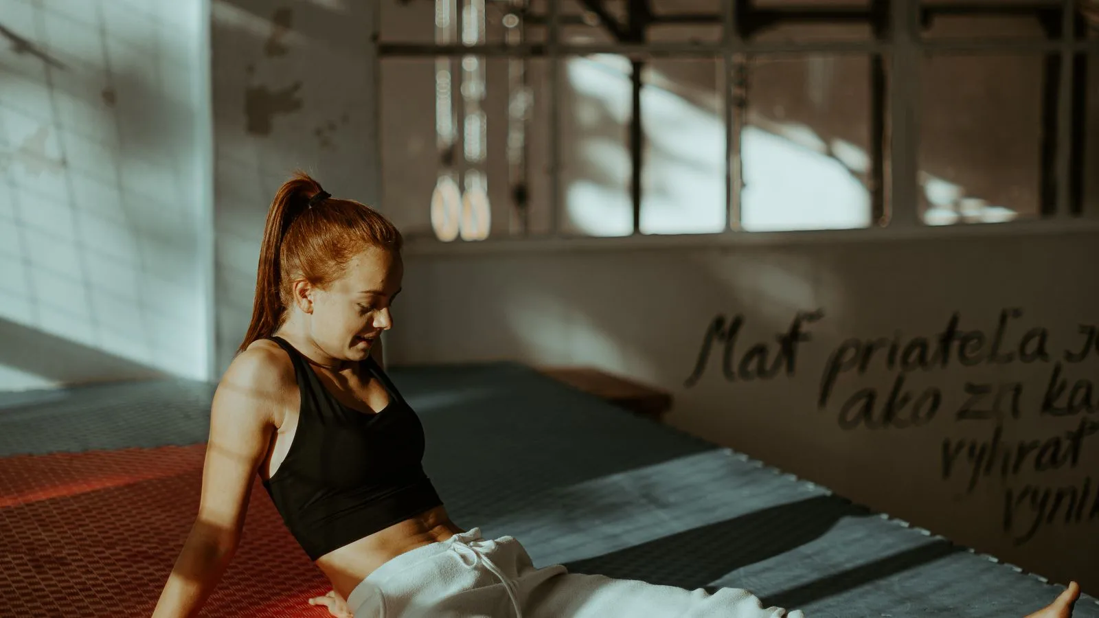 An athlete sitting on a gym mat in low afternoon light, looking down and thinking.